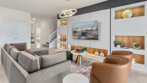 A modern living room with a grey sectional sofa, a brown armchair, and a round coffee table. A flat-screen TV is mounted on the wall above a wooden accent wall. - Ultimate 10 Bed Luxury Retreat With Epic Playroom (Clermont)