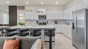 A modern kitchen with white cabinets, a stainless steel refrigerator, a central island with bar stools, and a dining area with a sofa and table. - Ultimate 10 Bed Luxury Retreat With Epic Playroom (Clermont)