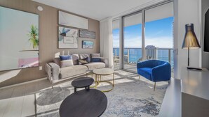 A modern living room with a sofa, coffee table, and blue armchair, large windows with a city view, and artwork on the walls.