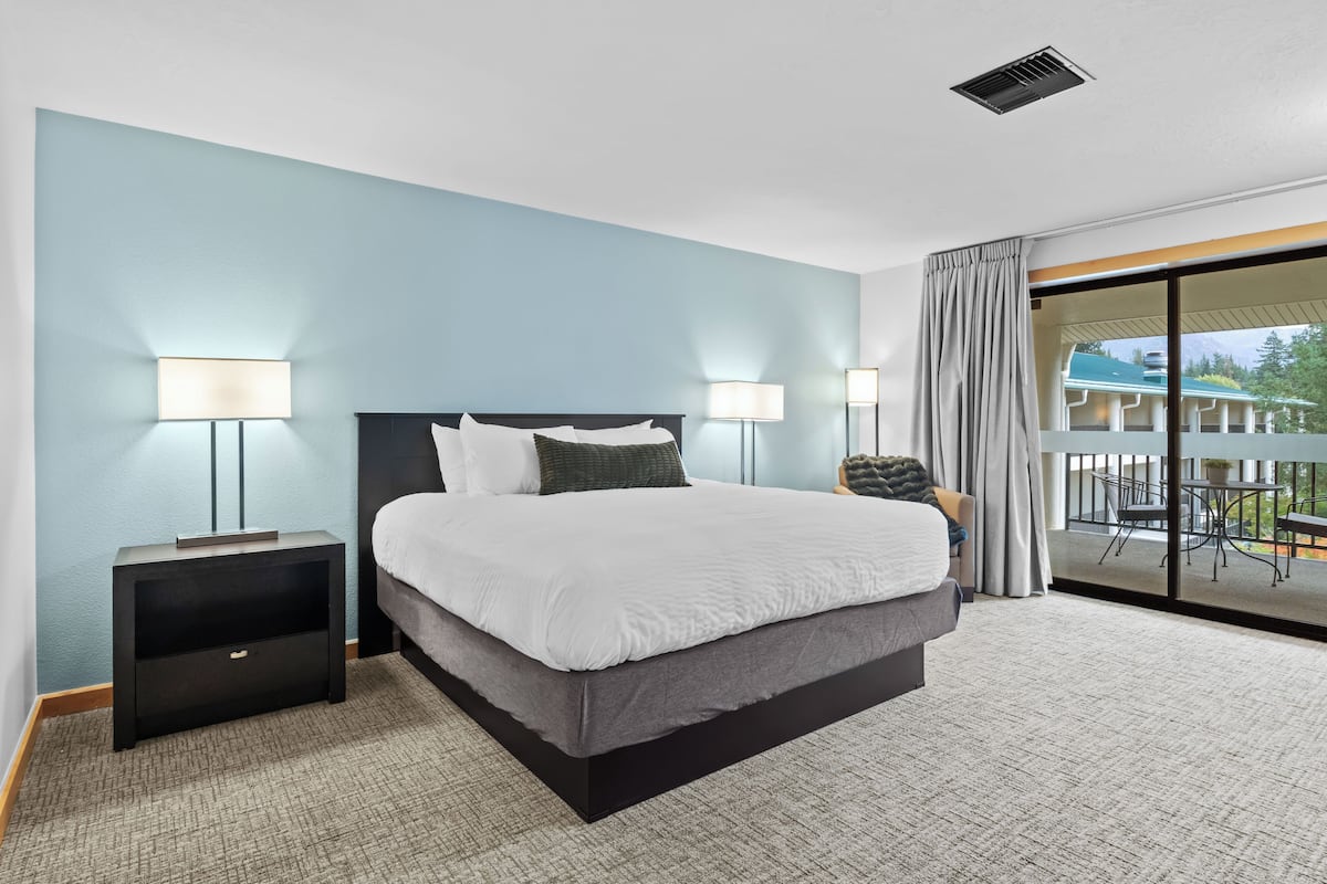 Une chambre d’hôtel moderne avec un grand lit, deux lampes de chevet, une petite table de nuit et une vue sur l’extérieur grâce à une porte-fenêtre coulissante.