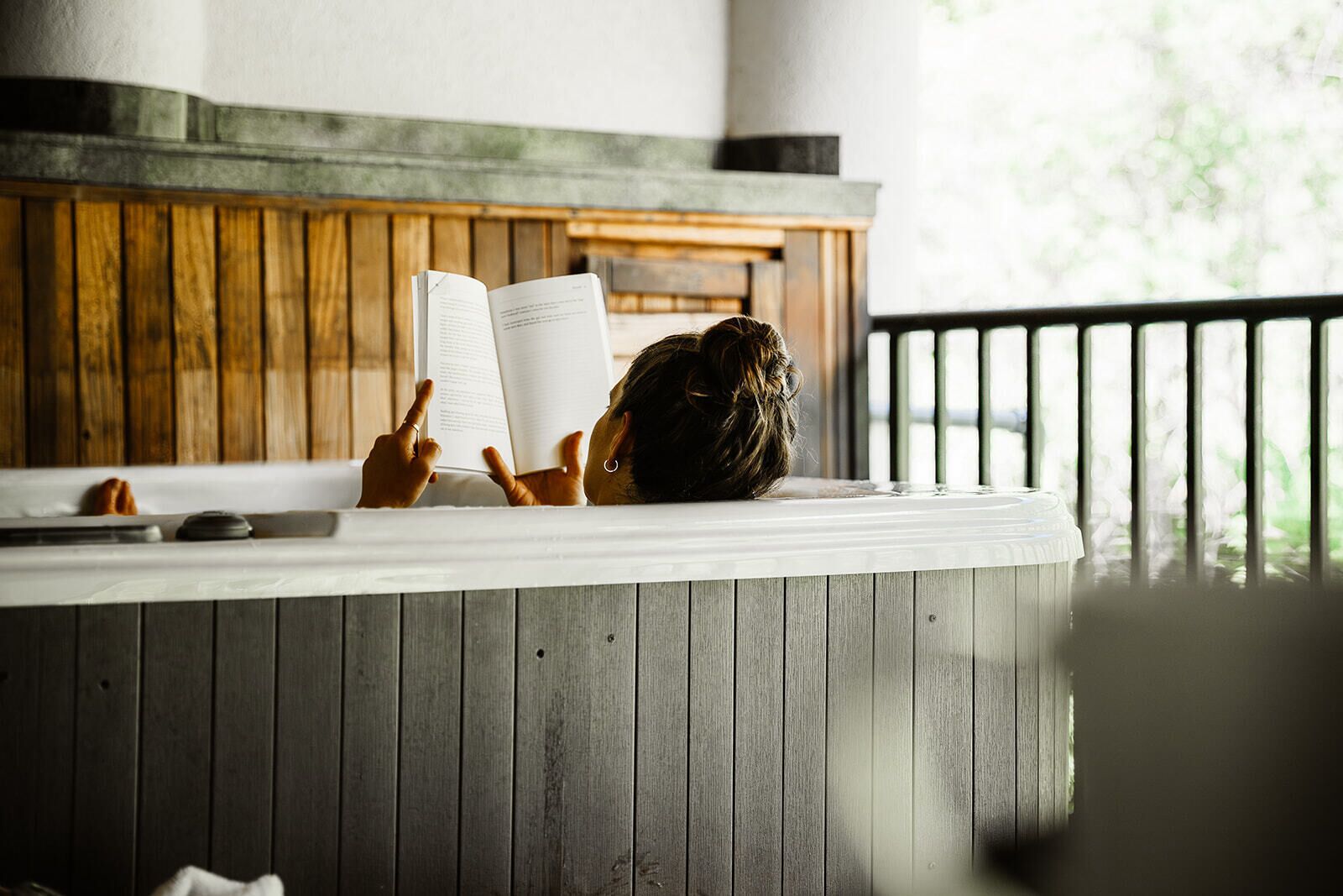 A person reading a book in a room with wooden paneling and a window with blinds.