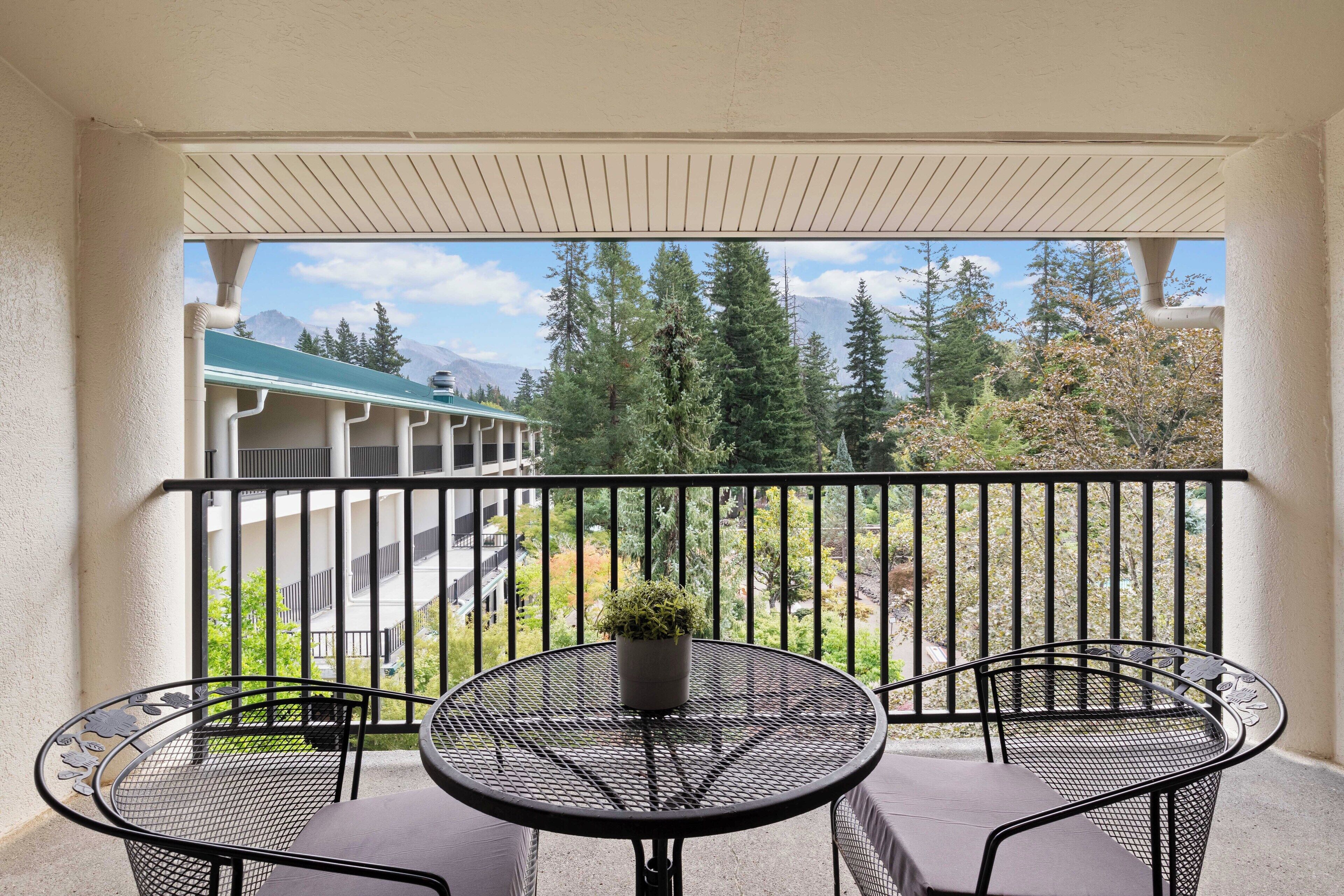 A balcony with a table and two chairs, overlooking a building with a green roof and trees.
