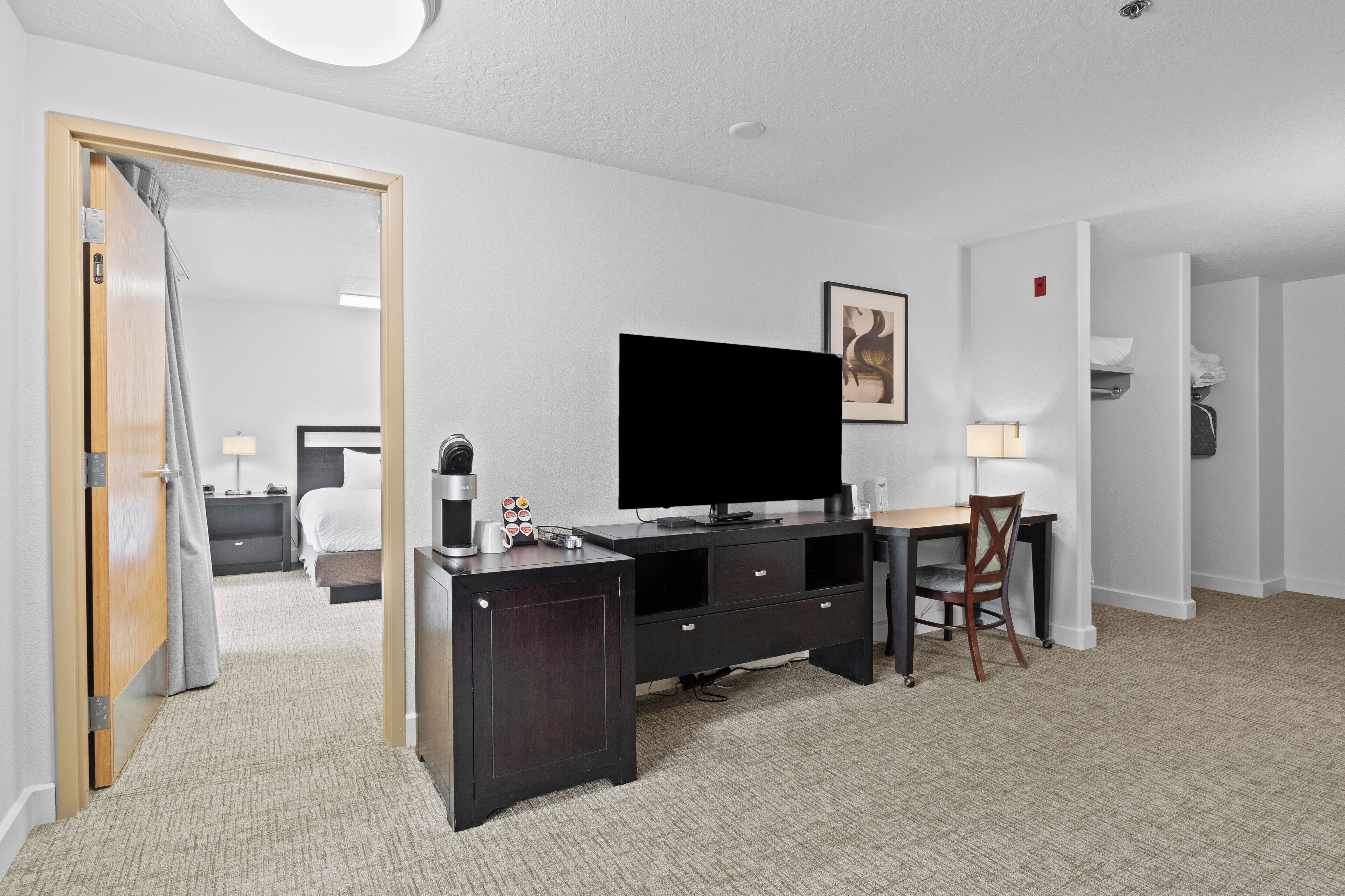 A hotel room with a large flat-screen TV, a wooden desk, a chair, a bed, and a bathroom visible through an open door.