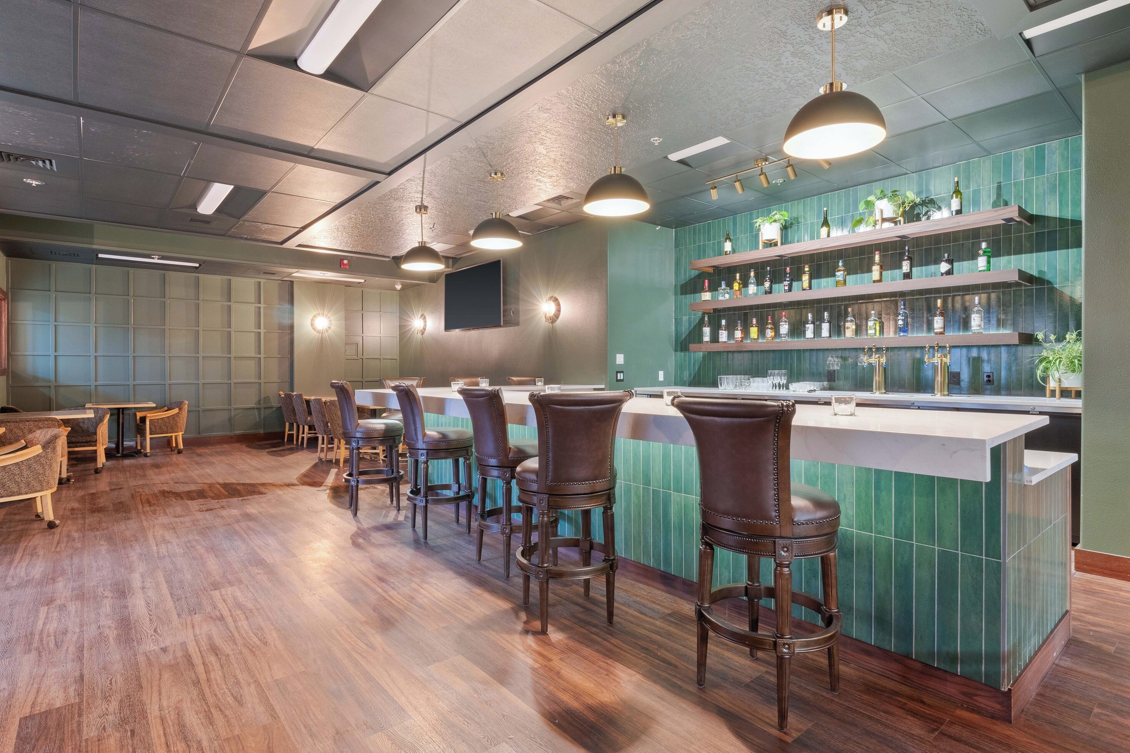 Eine Bar mit Holzboden, Barhocker, einer Theke mit gröner Fliesenwand und Deckenleuchten.