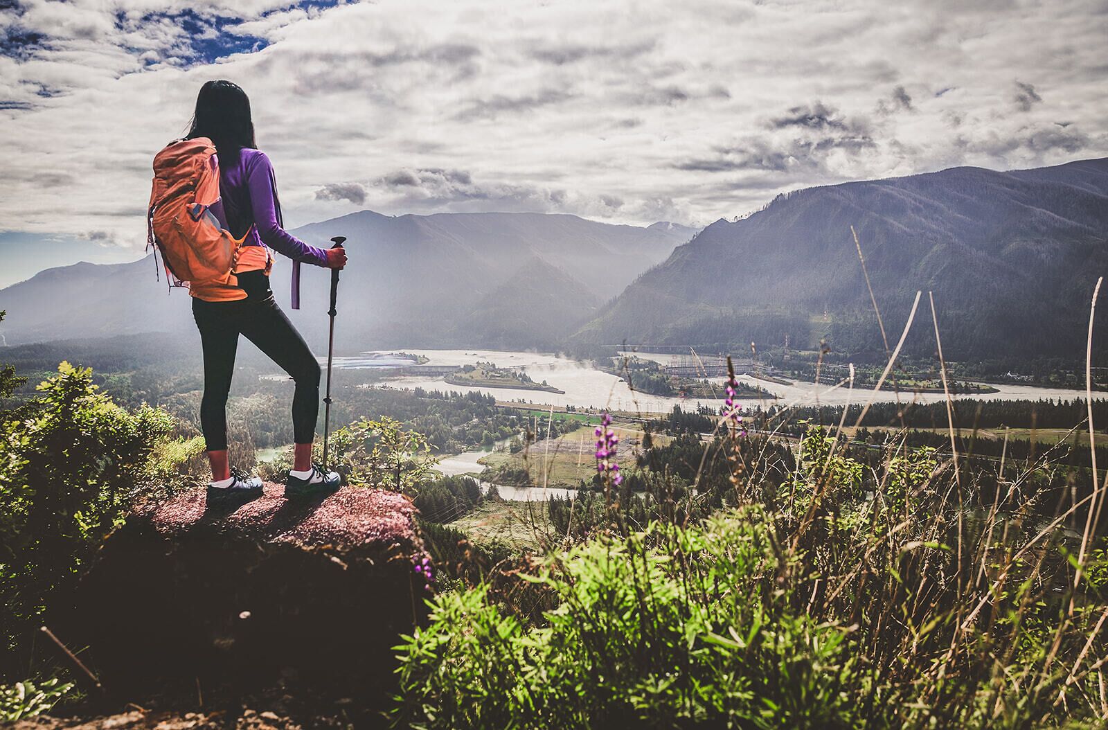 Una persona haciendo senderismo con una mochila y bastones de trekking, observando un valle con montañas y un río.