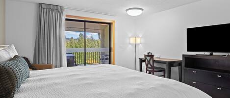 A hotel room with a large bed, a desk, a chair, a television, and a balcony with a view of trees.