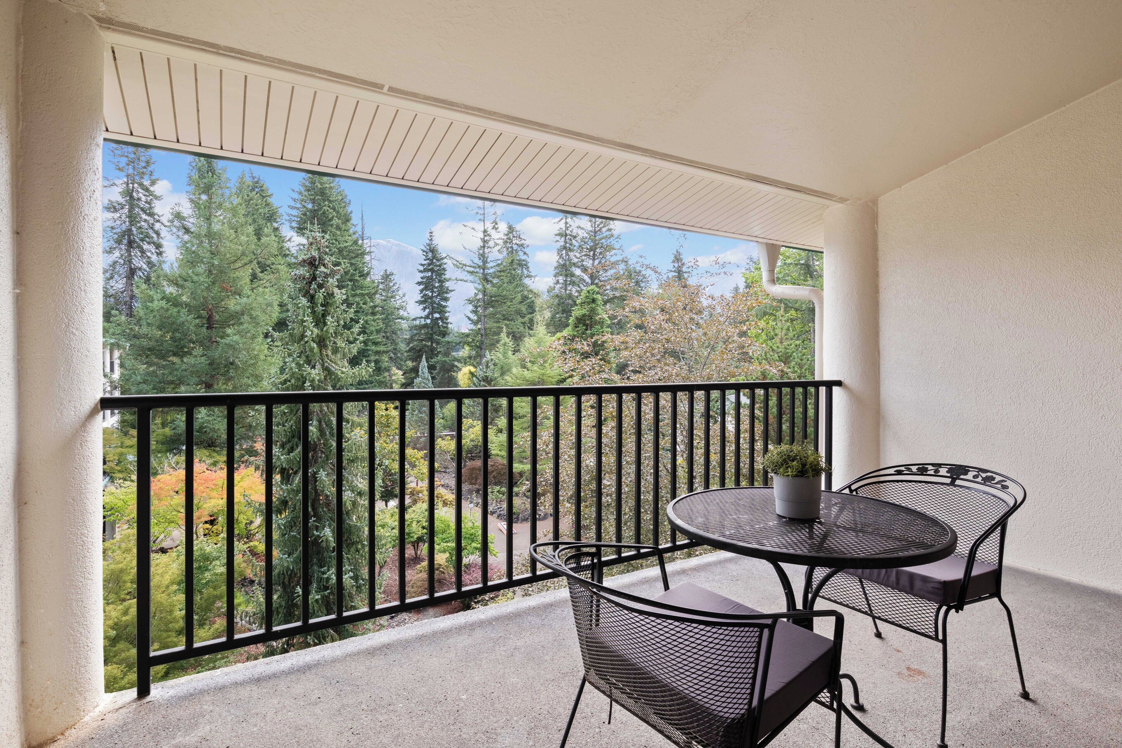 A balcony with a round table and two chairs, a potted plant, and a view of a forested area.