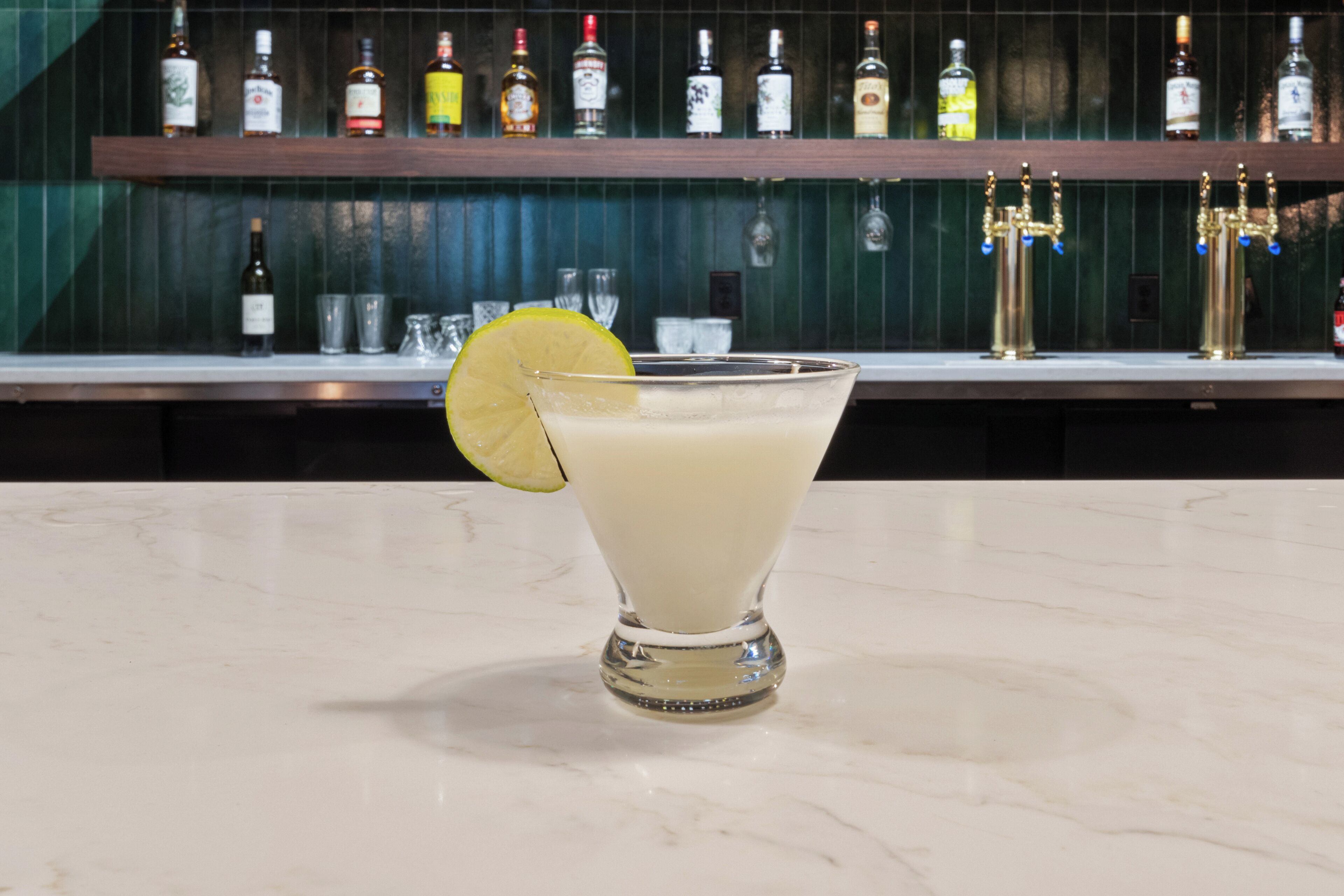 A bar area with a glass of a light-colored drink garnished with a lime slice, a selection of bottles on shelves, and a bar counter.