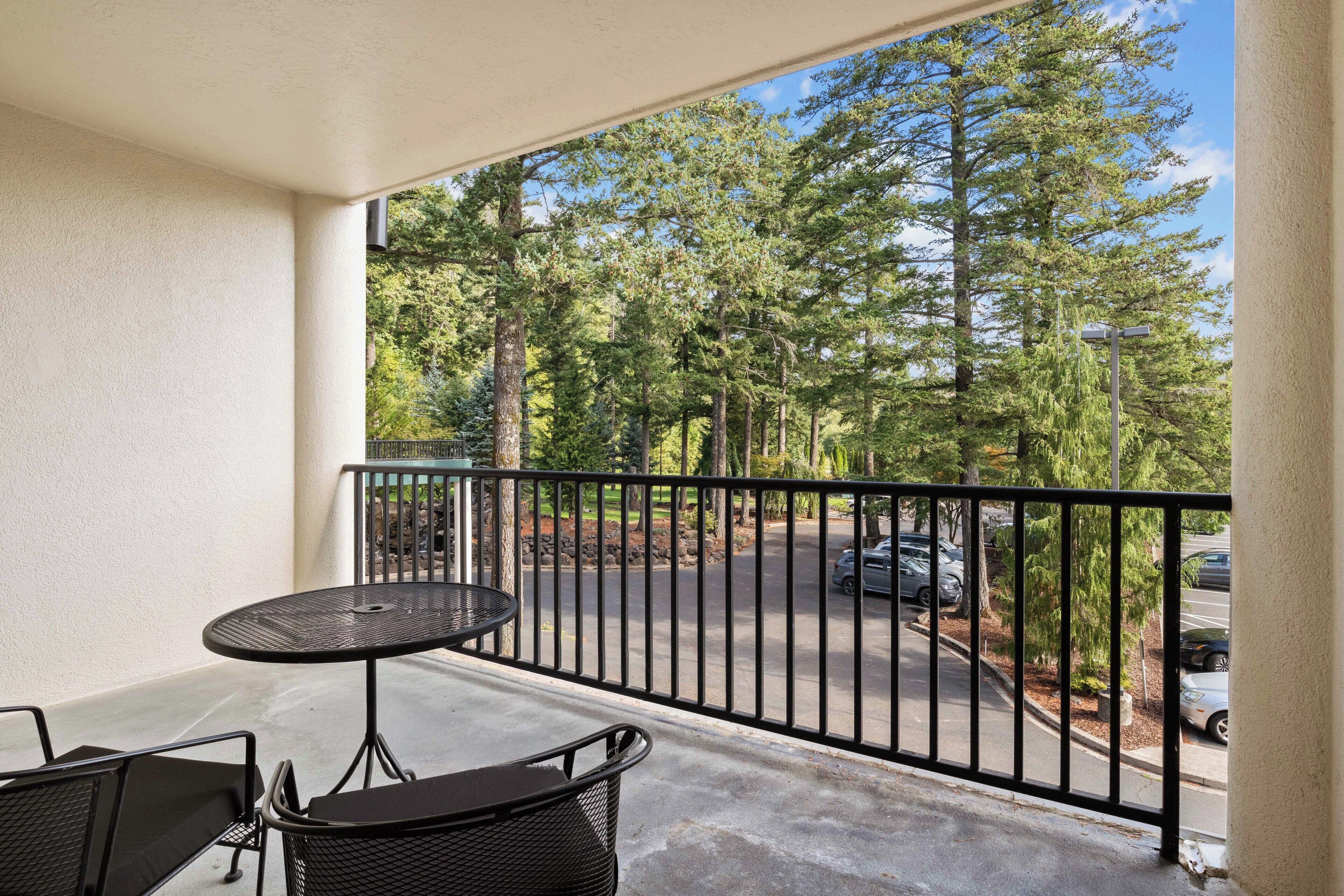 A balcony with a round table and two chairs, overlooking a street with parked cars and a row of trees.