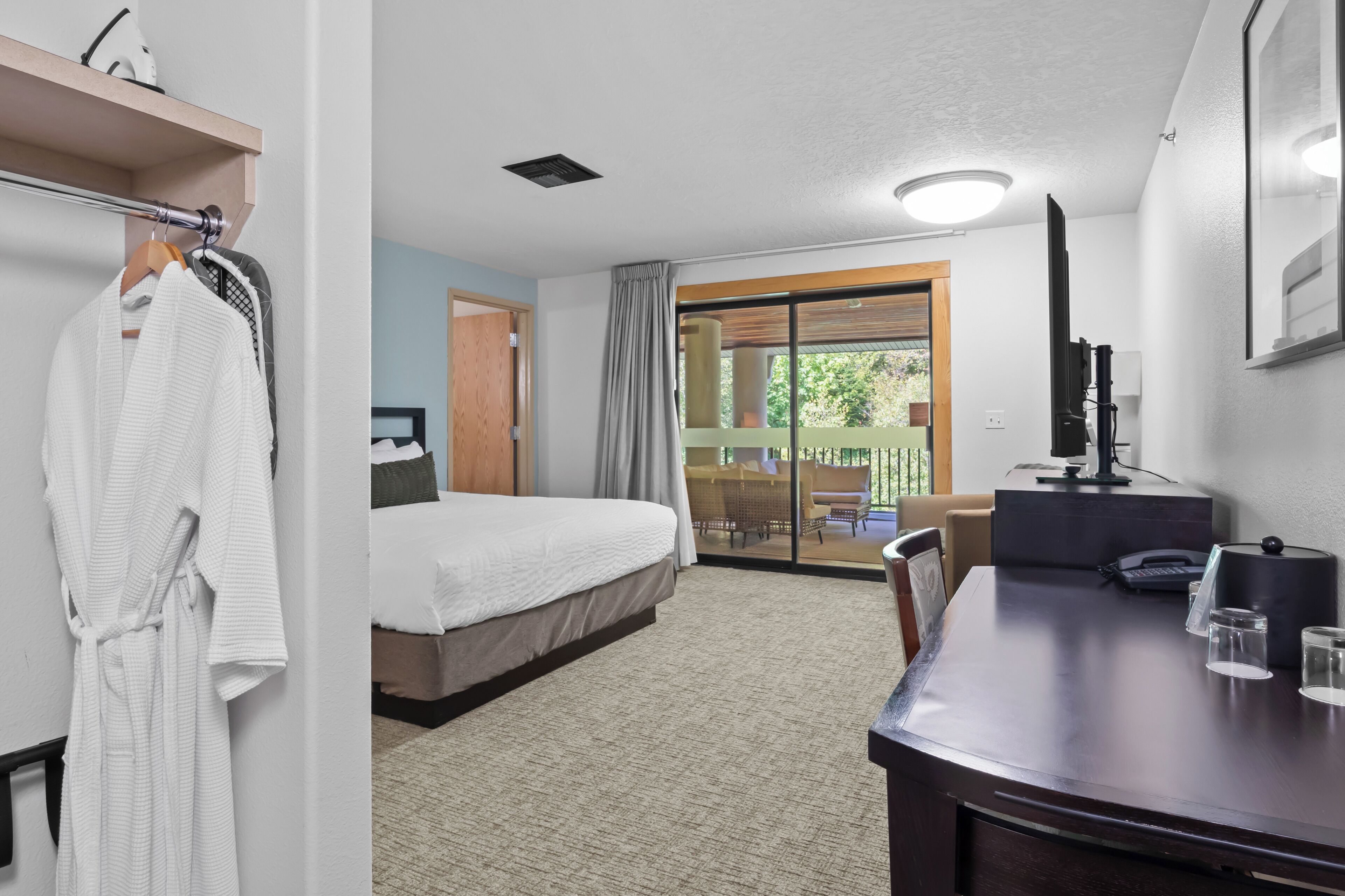A hotel room with a bed, a desk with a computer, a TV, and a balcony with a view of greenery.