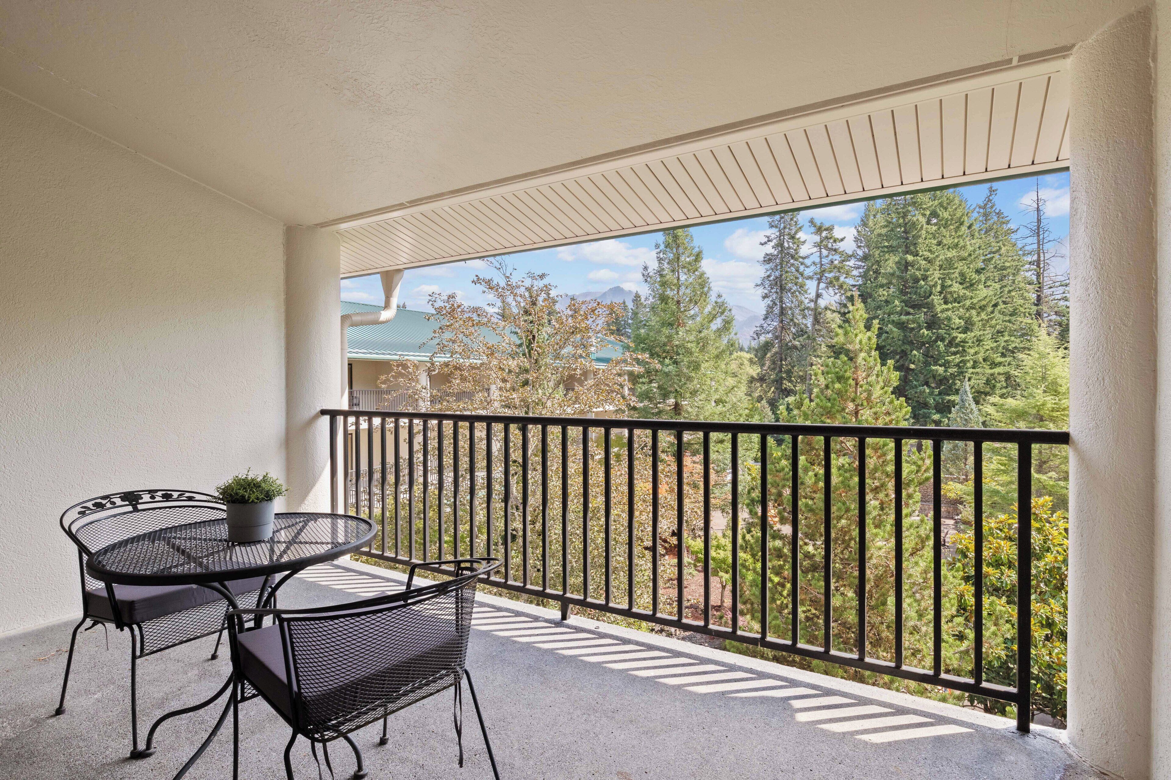 Un balcon avec une table ronde et des chaises, un pot de fleurs et une vue sur les arbres et les montagnes.
