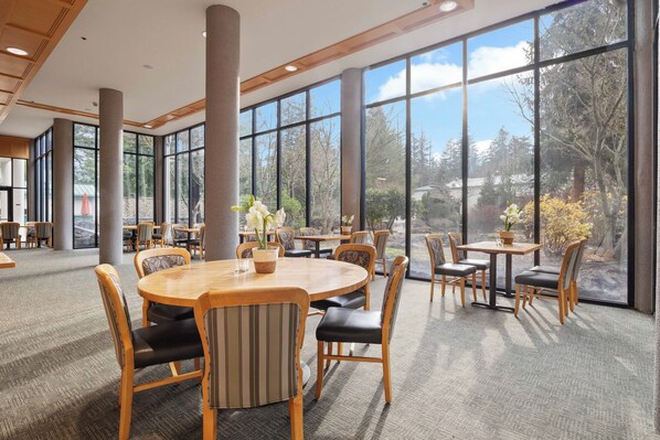 A spacious indoor area with large windows, wooden tables, and chairs, providing a view of the outdoors.