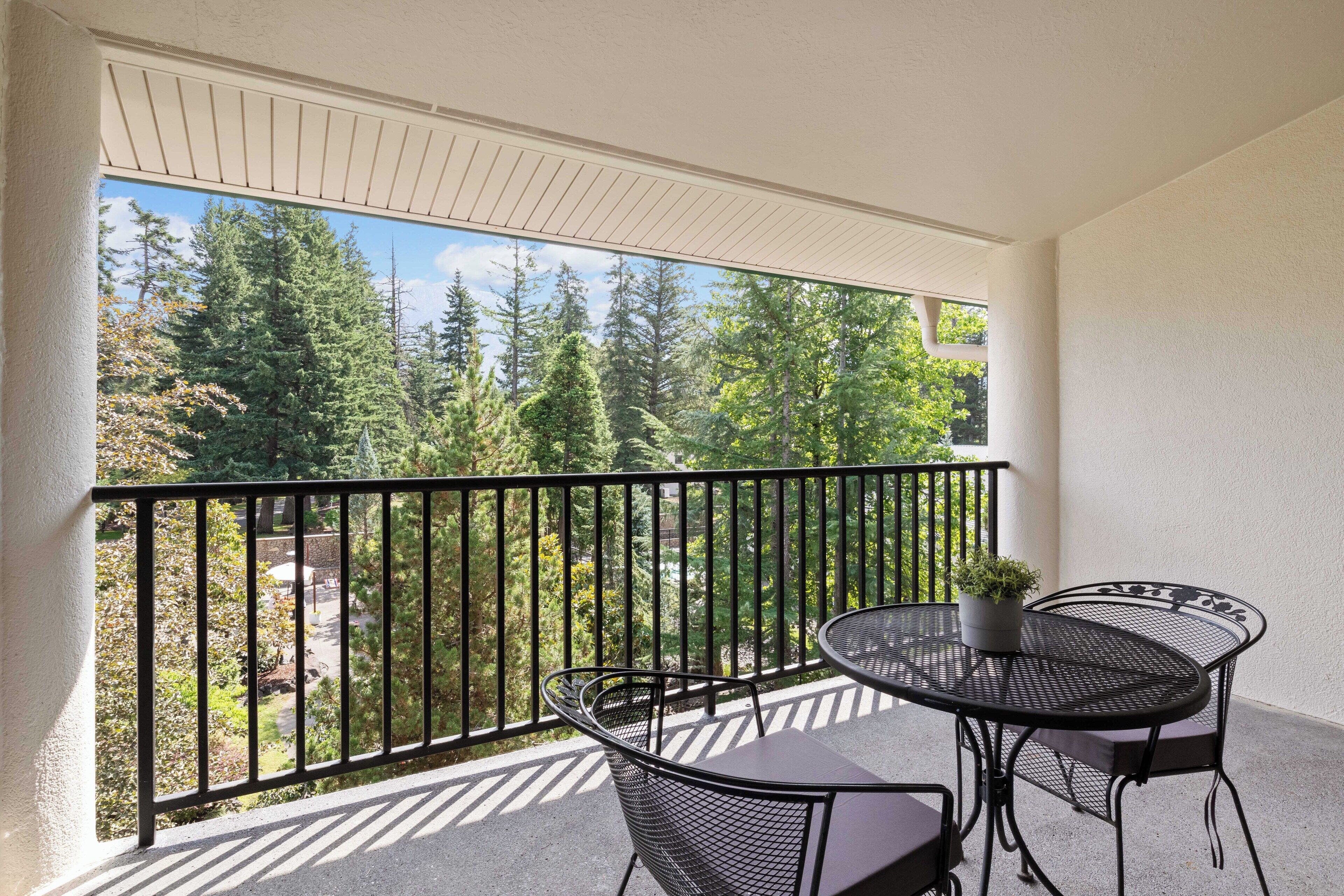 A balcony with a round table and two chairs, a potted plant, and a view of trees and a building.