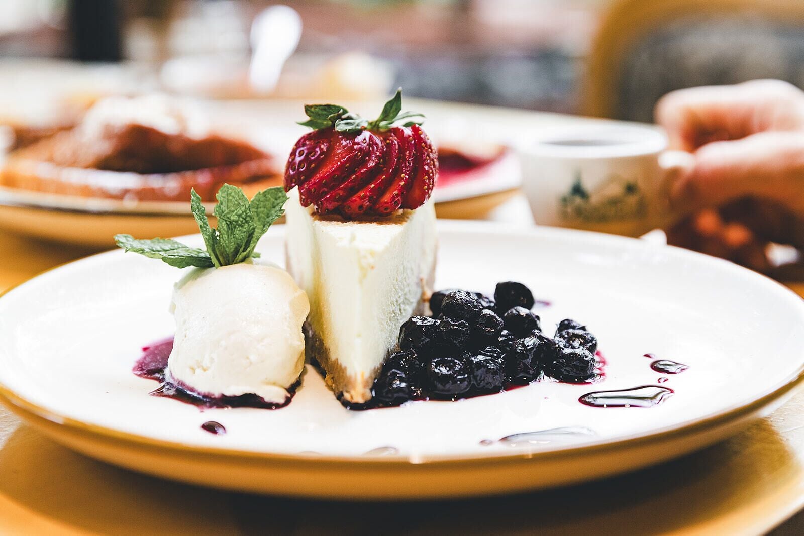 Un plato de postre con una porción de pastel de queso coronado con una fresa, una bola de helado de vainilla y arándanos.