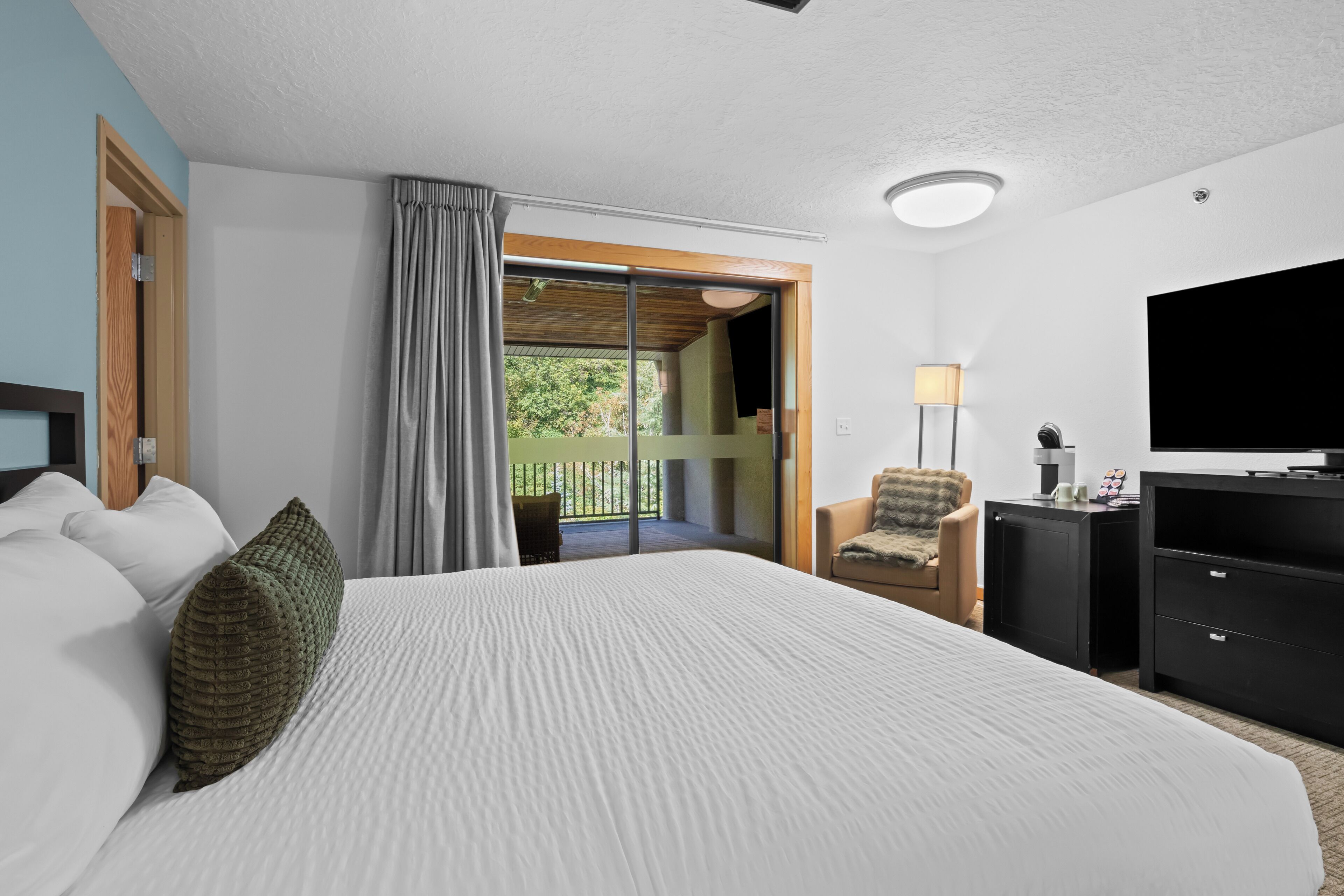 Habitación de hotel con una cama grande, televisión, sillón y balcón con vistas a la vegetación.