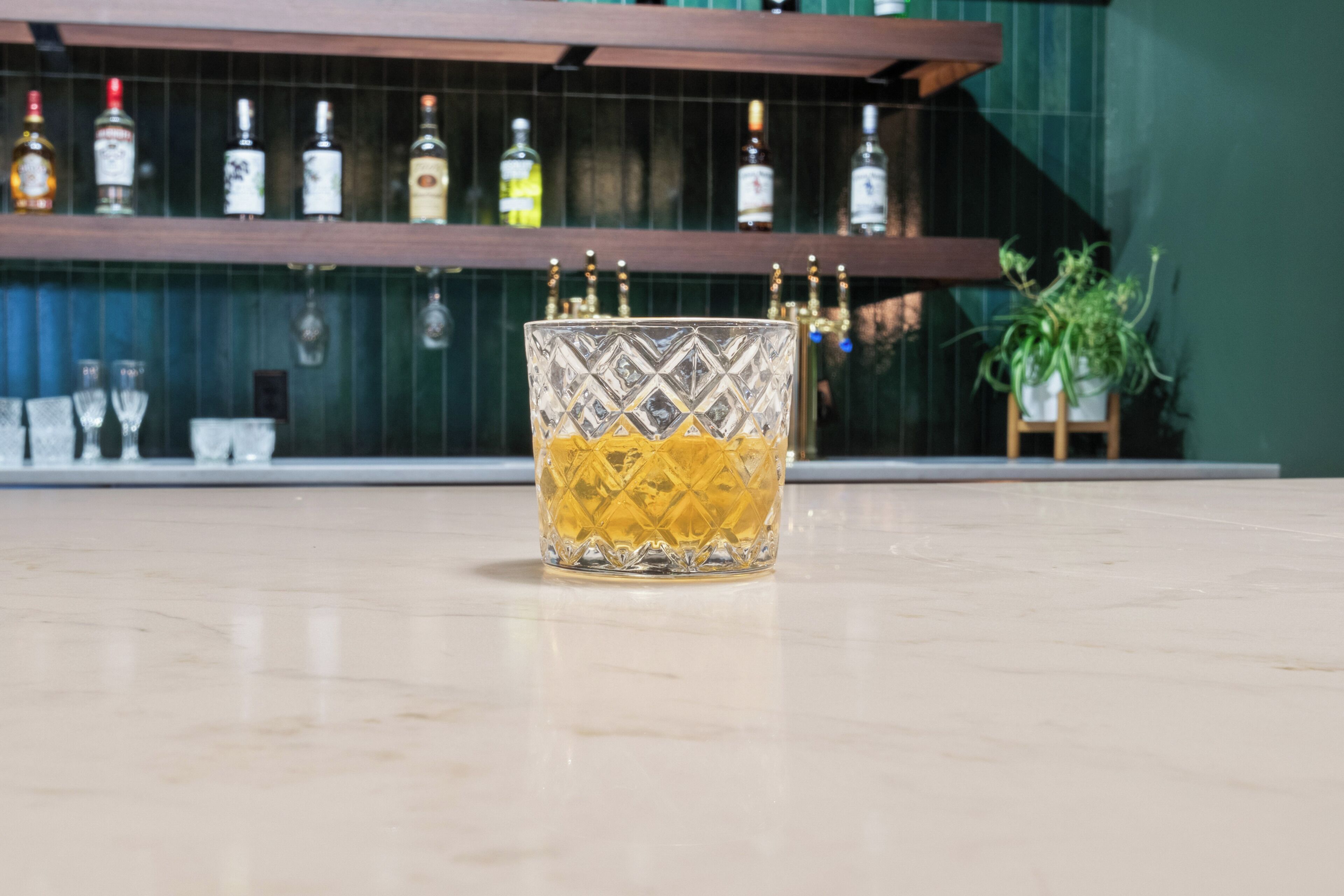 A bar area with a glass of amber-colored liquor, shelves with various bottles, and a potted plant.