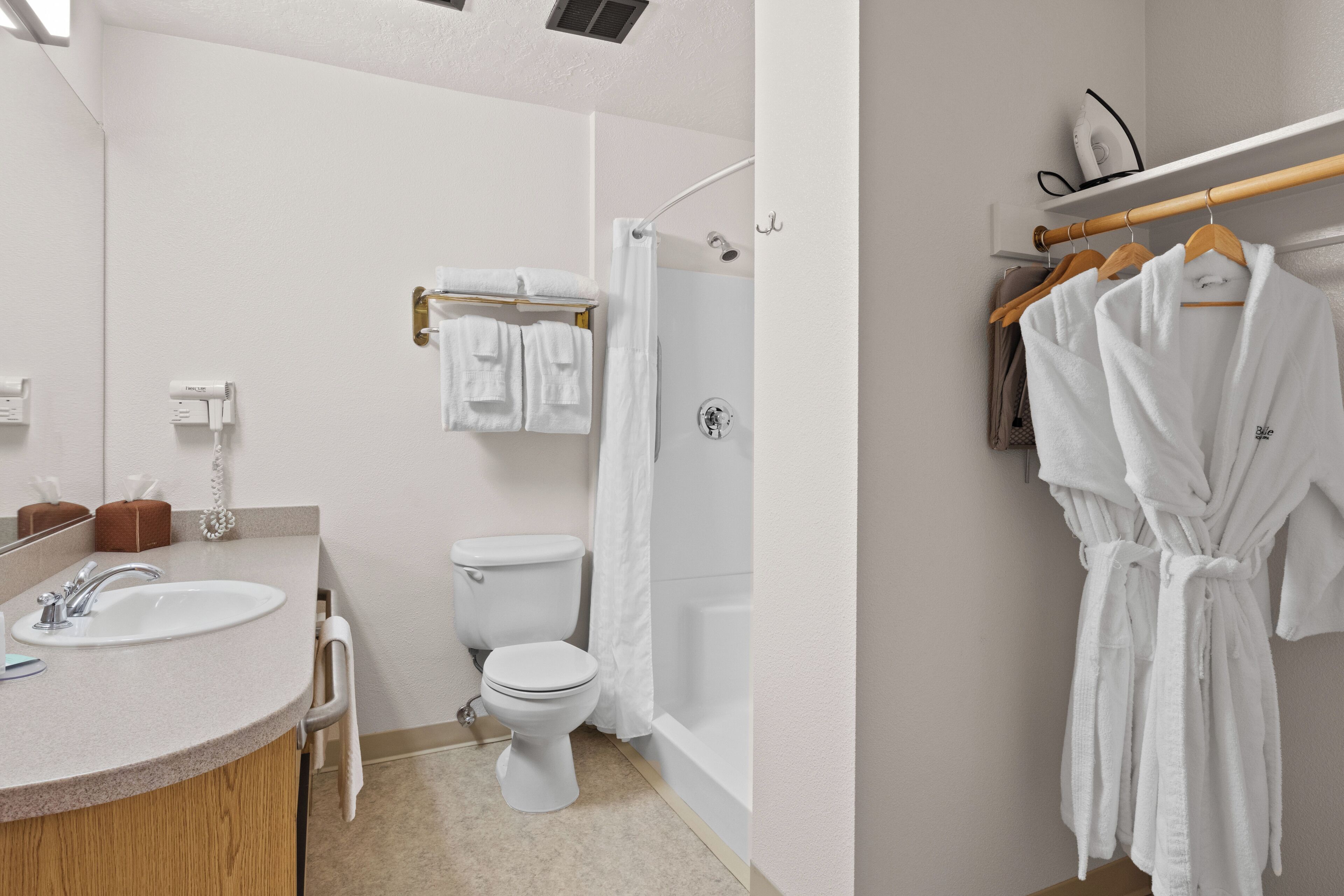 A bathroom with a sink, toilet, shower, and a rack with towels and robes.