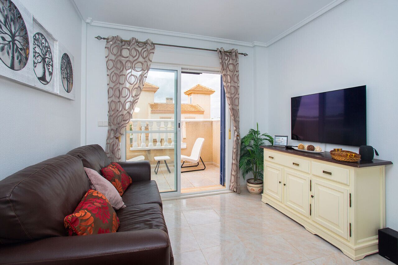 A living room with a brown leather sofa, a cream-colored cabinet, a flat-screen TV, and a sliding glass door leading to a balcony with a white chair and a potted plant.