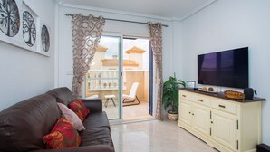 A living room with a brown leather sofa, a cream-colored cabinet, a flat-screen TV, and a sliding glass door leading to a balcony with a white chair and a potted plant.