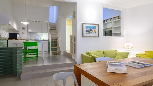A modern kitchen with white cabinets, a dining area with a wooden table and chairs, and a living area with a green sofa and a bookshelf. - Apartments Florence-San Lorenzo Market (Florence)
