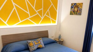 Une chambre d’hôtel avec un lit, une tête de lit, une œuvre d’art murale, une petite table et un rideau bleu.