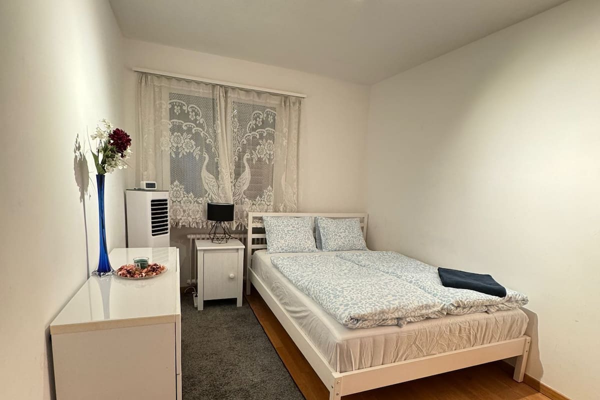 Un dormitorio con cama, mesitas de noche, cómoda, ventana con cortinas de encaje y lámpara en el techo.