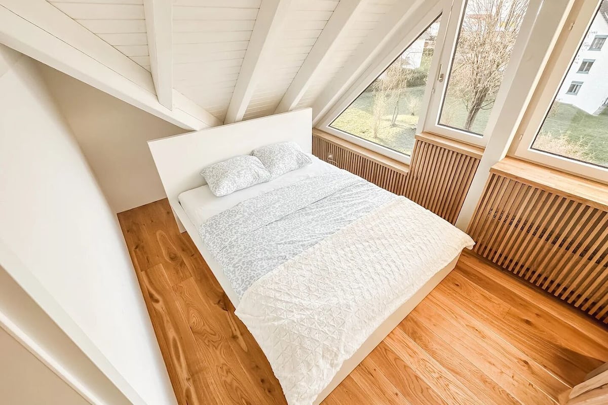 Un dormitorio con techo inclinado, una cama con ropa de cama estampada y ventanas amplias que ofrecen vistas al exterior.