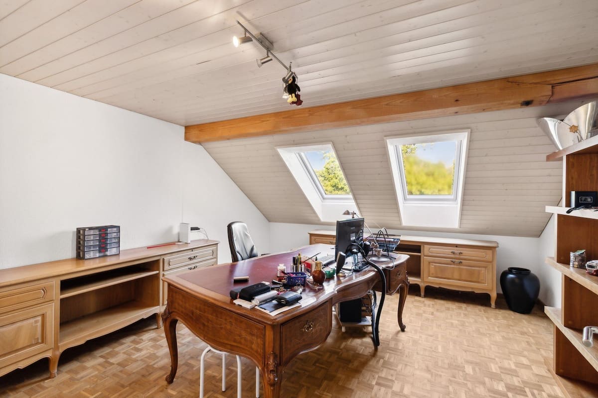 Amplia habitación en ático con escritorio de madera, computadora y silla. Dos ventanas permiten la entrada de luz natural.
