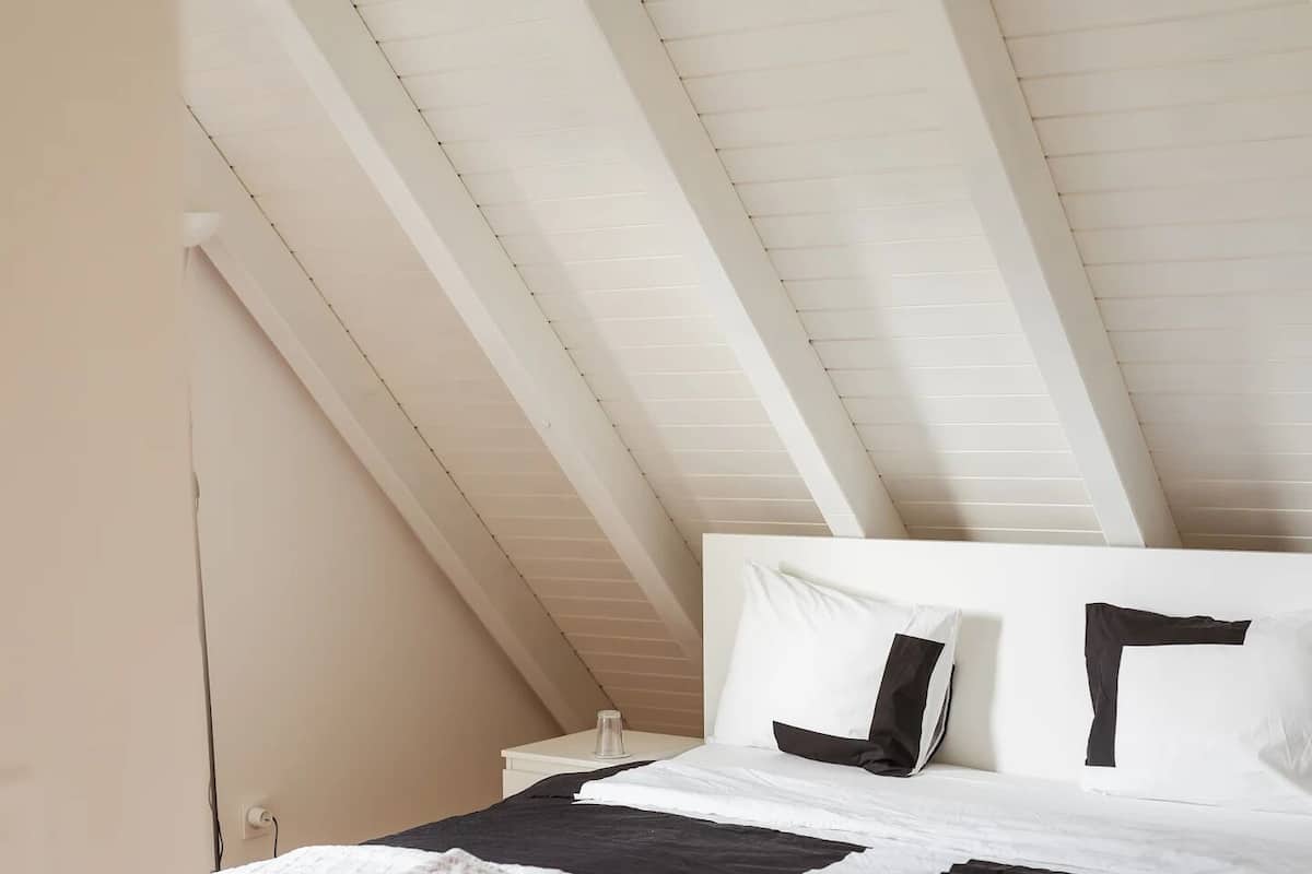Un dormitorio con techo inclinado, cama con ropa de cama blanca y negra, y piso de madera.