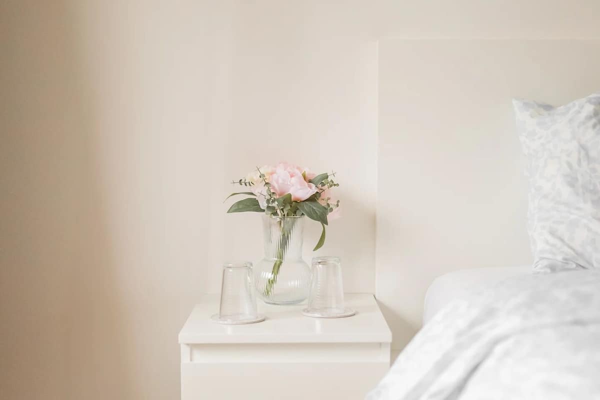 Un dormitorio con una mesita de noche blanca, un jarrón con flores y una cama con edredón blanco.