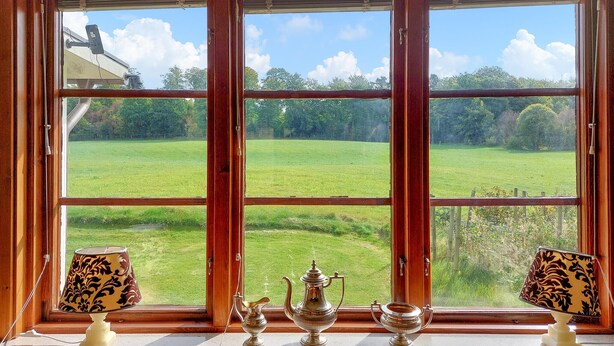 Ein Zimmer mit großem Fenster und Blick auf ein grünes Feld und Bäume. Zwei Lampen stehen auf dem Fensterbrett.