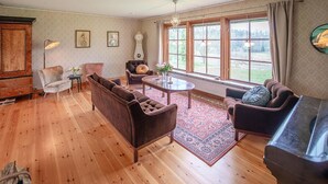 Ein geräumiges Wohnzimmer mit Holzböden, einem braunen Sofa, einem kleineren Sessel, einem Couchtisch und einem Klavier. Große Fenster mit Vorhängen, eine Kristallleuchter und gerahmte Bilder schmücken die Wände.
