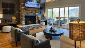 A modern living room with a stone fireplace, a flat-screen TV, and a large window offering a view of the outdoors. - 5 mi. to Asheville•HotTub•Sauna•MtnViews•FirePit•PoolTable (Swannanoa)