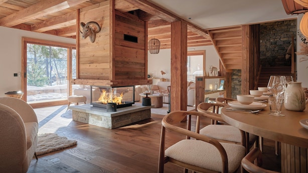 Un intérieur chaleureux en bois, avec une cheminée, un manteau de cheminée en pierre et un coin repas comprenant des chaises et une table.