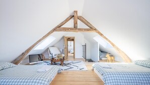 Un dormitorio acogedor en el ático, con techo inclinado, vigas de madera, dos camas con ropa de cama de cuadros, una mesita con un jarrón y vista a la habitación contigua.