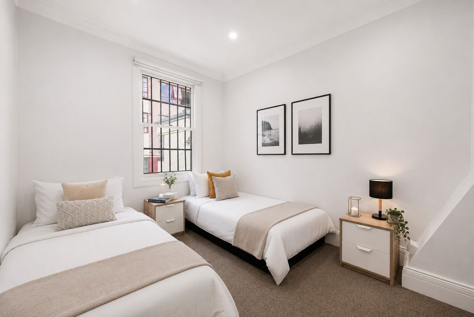 A bedroom with two beds, a window, a bedside table with a lamp, and two framed pictures on the wall.