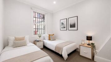 A bedroom with two beds, a window, a bedside table with a lamp, and two framed pictures on the wall.