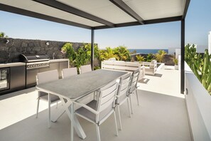 Een terras buiten met een barbecue, eettafel en stoelen, met uitzicht op de kust.