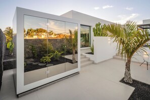 A modern building with a glass facade, a stone wall, and a small garden with plants and a palm tree. - AMBIANT by Buenavilla | Serene elegance in a privileged location (Puerto Calero)