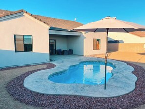 A swimming pool with a curved edge, surrounded by a pebble border, and a patio area with outdoor furniture. - New build! Pool | 3bd | 2ba | Sleeps 7 | Views (Fort Mohave)