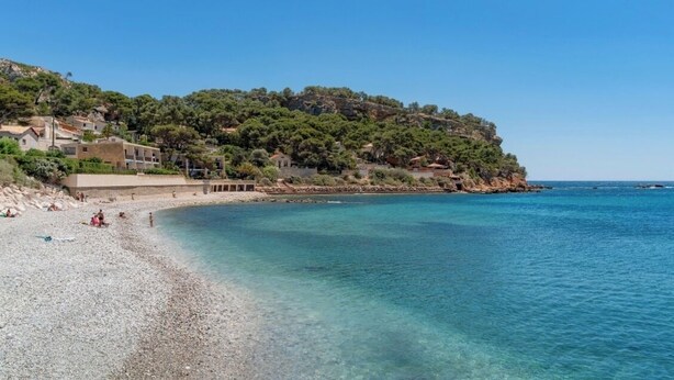 A coastal beach with clear water, a pebble beach, and buildings on a hillside.