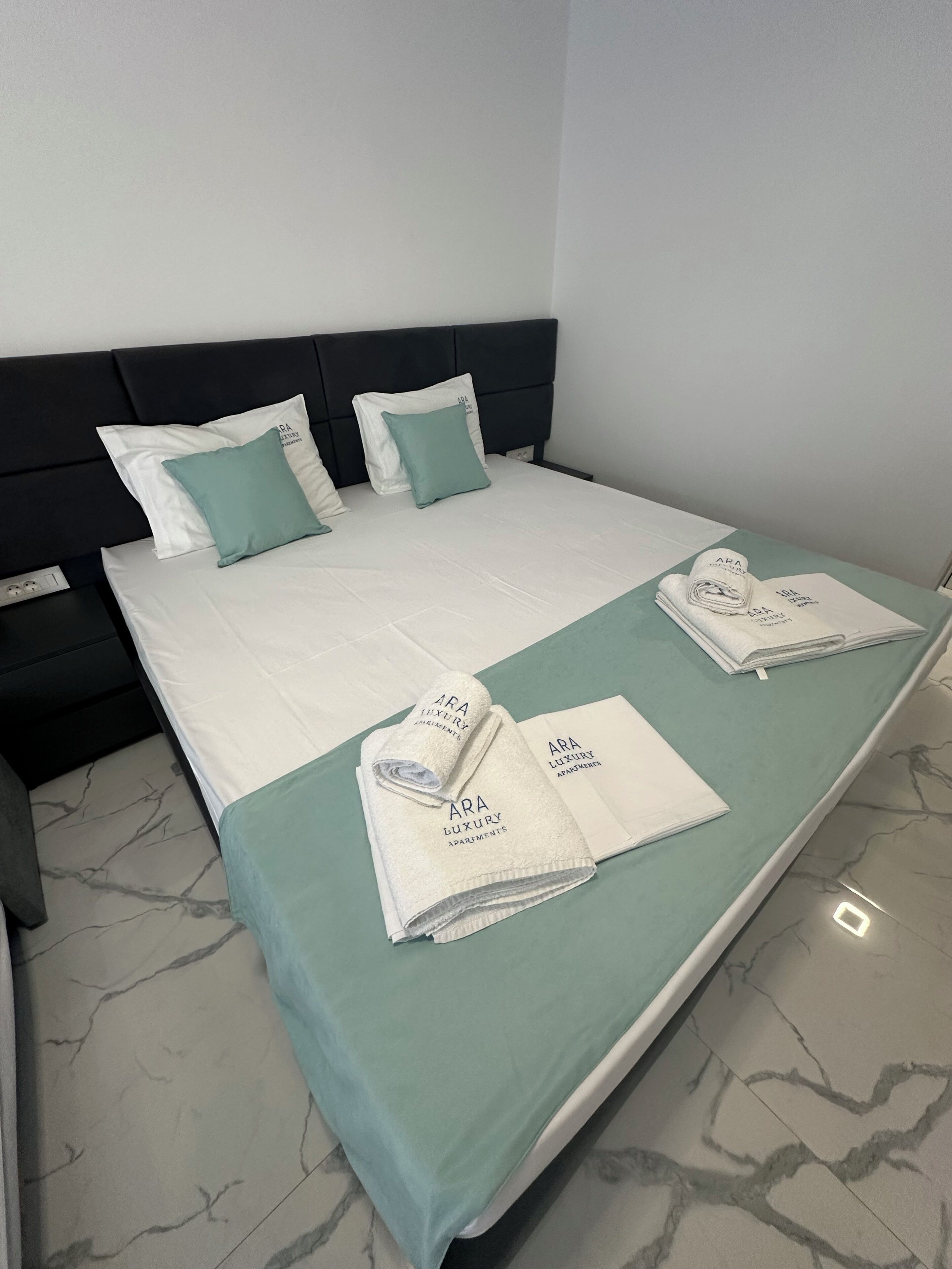 A neatly made bed with a green bedspread, white pillows, and a folded towel with "ARA Luxury Apartments" branding.