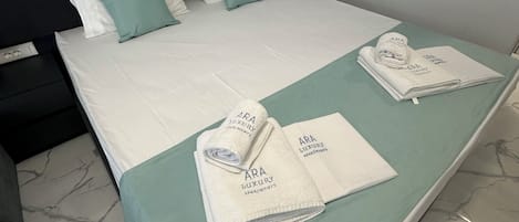 A neatly made bed with a green bedspread, white pillows, and a folded towel with "ARA Luxury Apartments" branding.