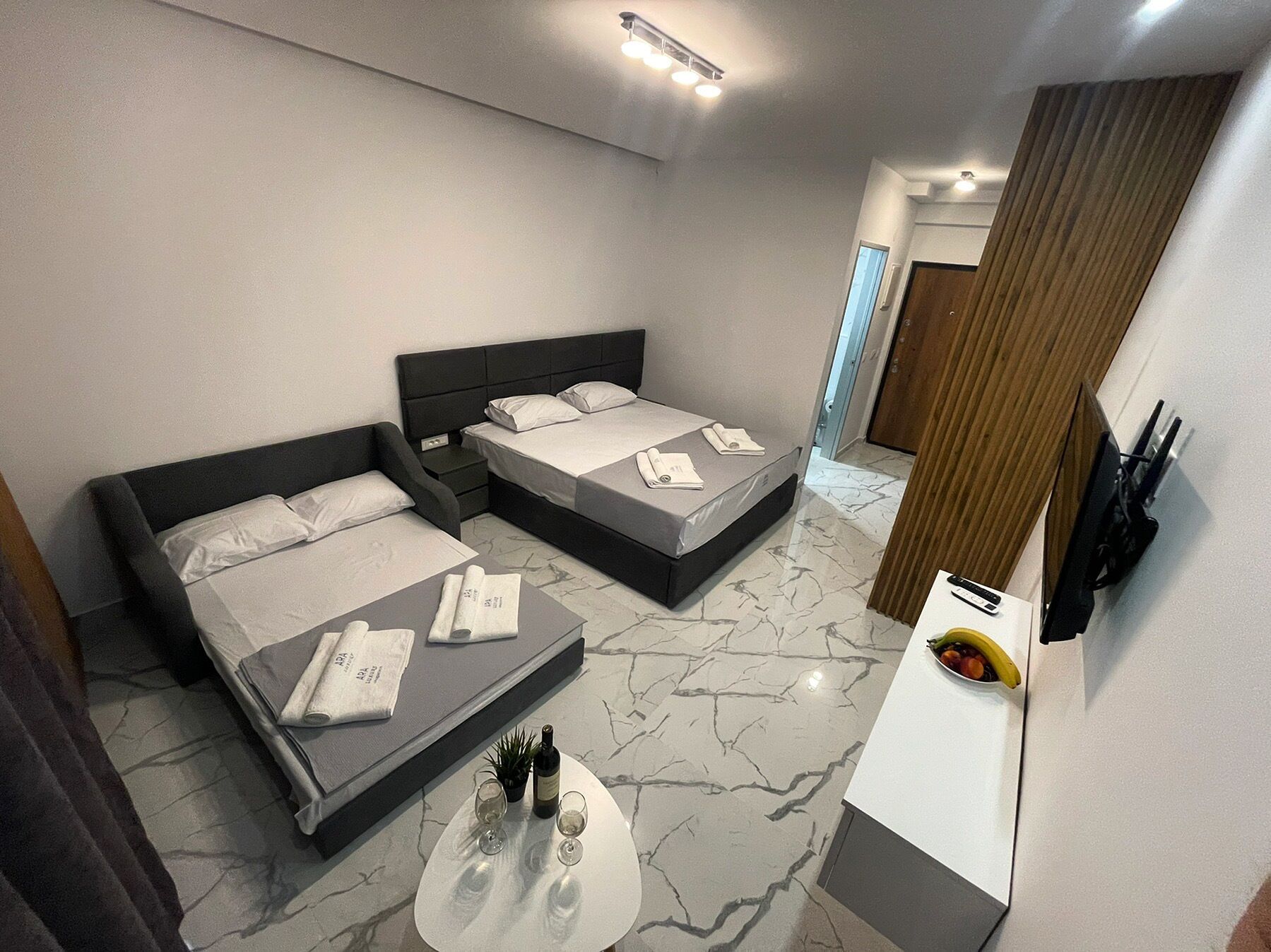 A modern hotel room with two beds, a small table with a bottle and glasses, and a TV on the wall.