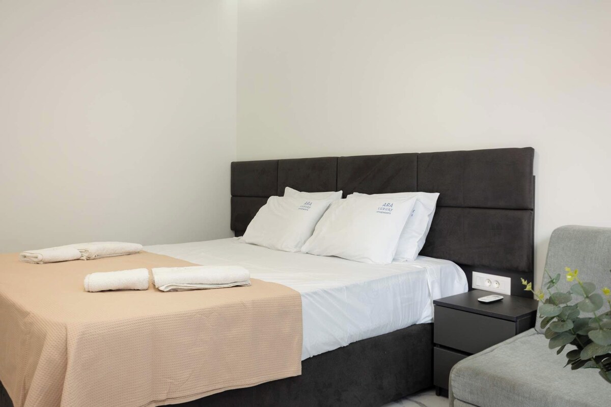 Una cama bien hecha con sábanas y almohadas blancas, una mesita de noche con lámpara y una pequeña planta en maceta.