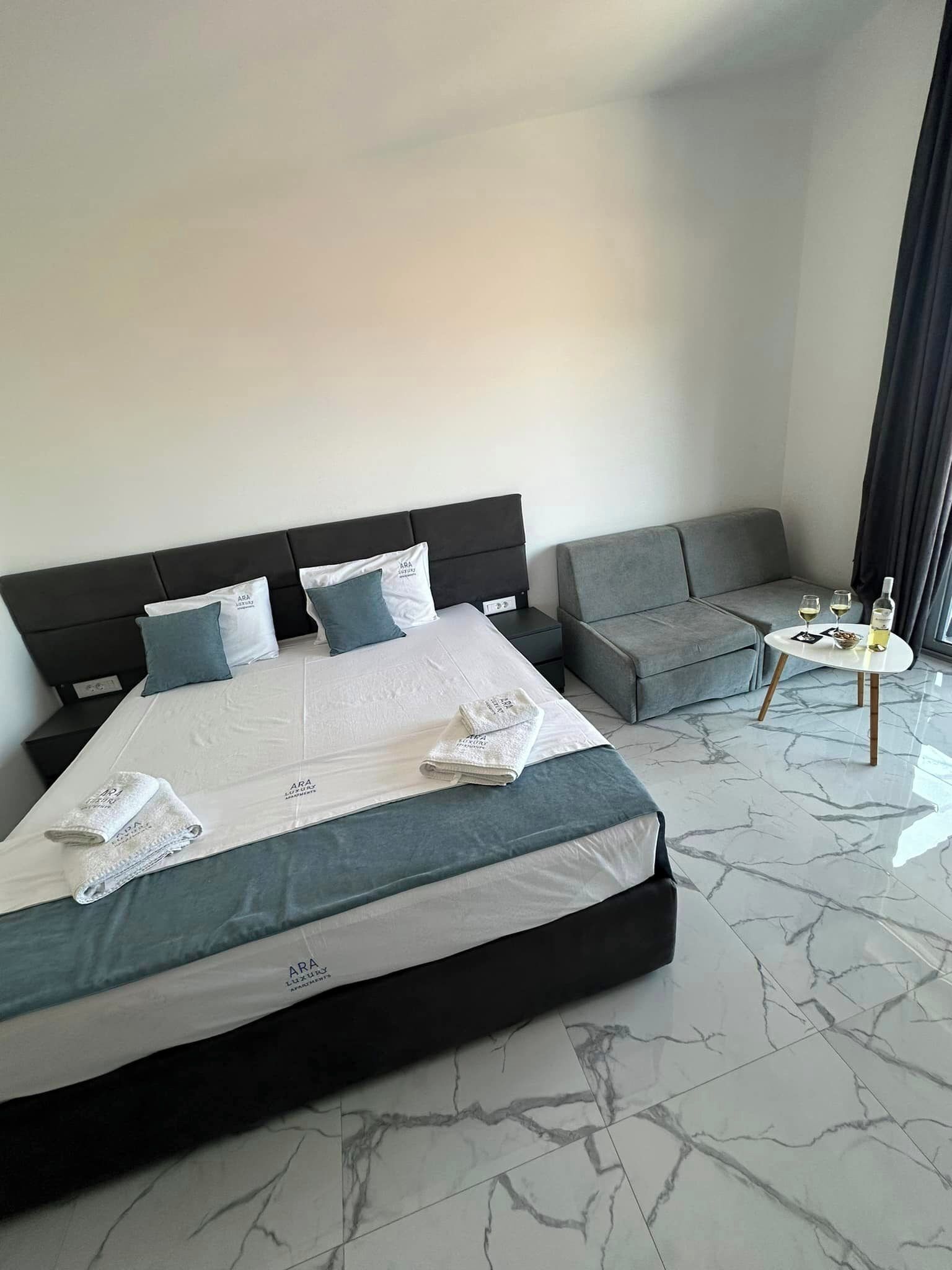 Ein modernes Hotelzimmer mit einem großen Bett, einem grauen Sofa und einem kleinen runden Tisch mit einer Flasche und Gläsern.