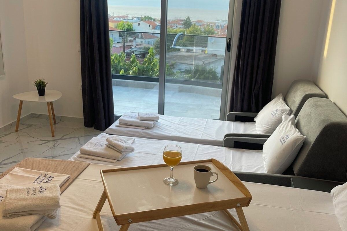 A hotel room with a bed, a small table with a glass of orange juice and a cup, a sofa, a balcony with a view of buildings, and an air conditioning unit.