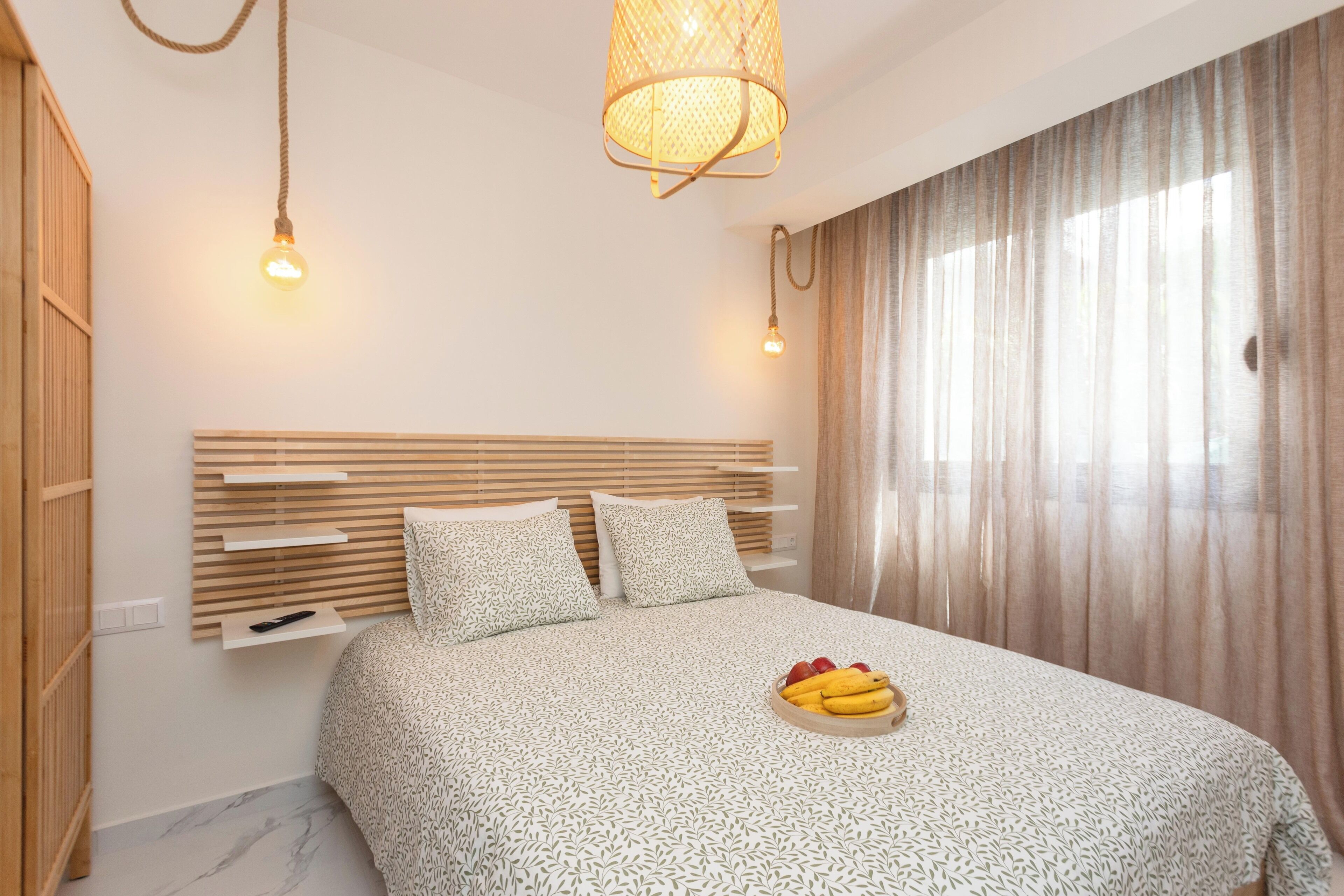 Um quarto moderno com cabeceira de madeira, cama com colcha estampada, mesa de cabeceira com tigela de frutas e luminária pendente.