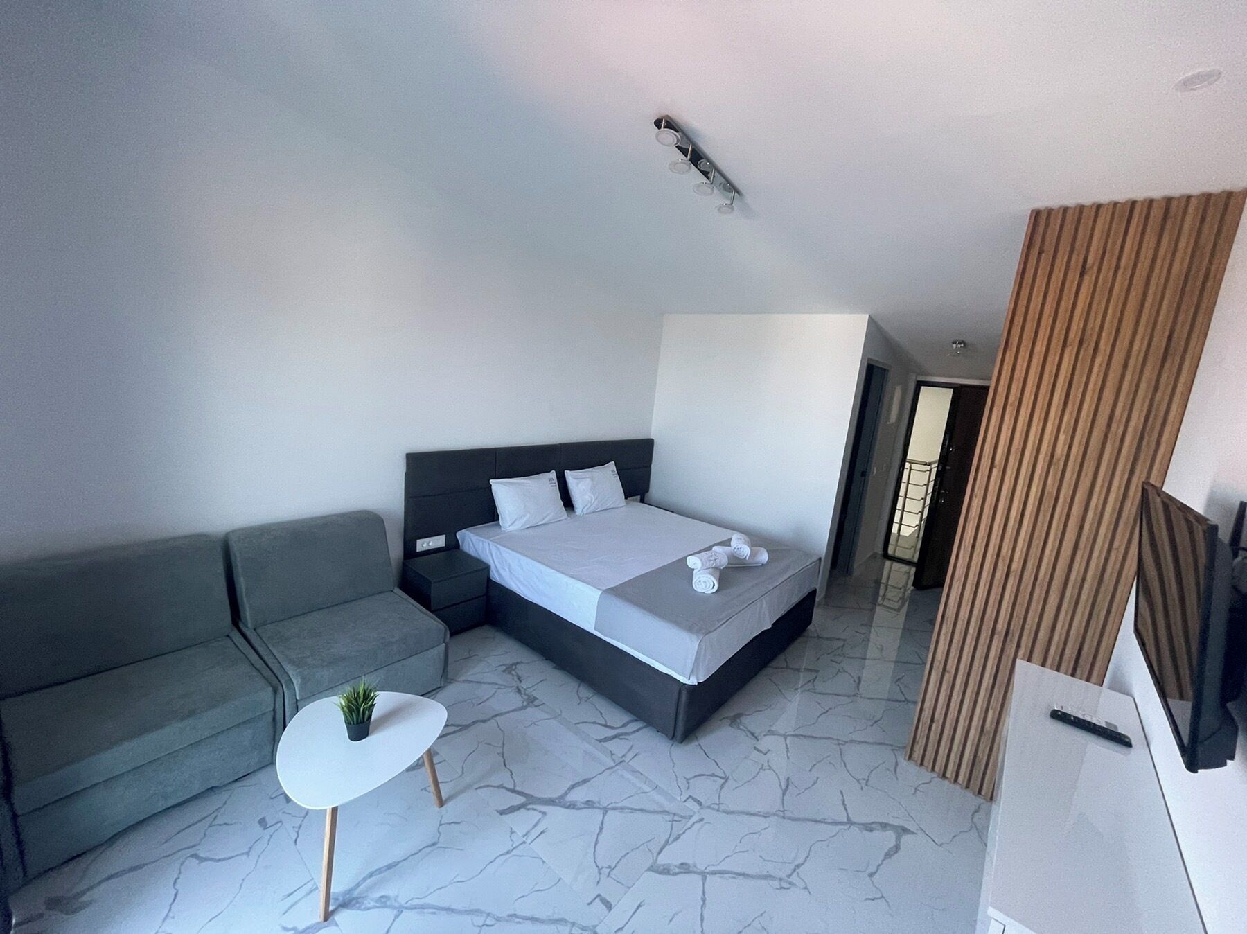 A modern bedroom with a bed, grey sofa, small white table, and a potted plant.