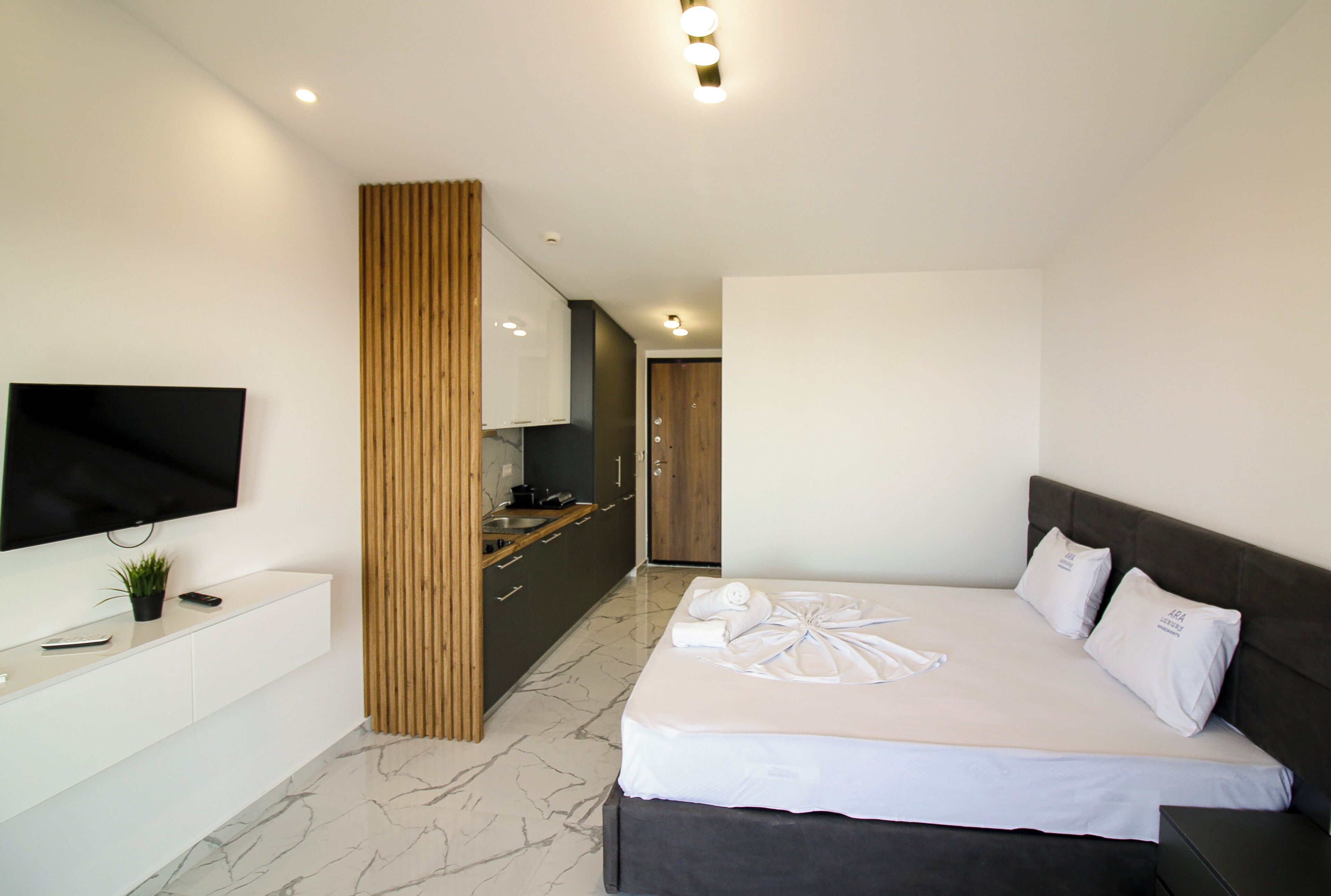 A modern hotel room with a large bed, a TV mounted on the wall, a wooden panel divider, and a small kitchenette area.